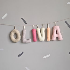 May include: A felt banner spelling "OLIVIA" in various pastel colors, including beige, lavender, pink, white, and peach. The letters are strung on a thin string against a gray wall decorated with scattered, elongated shapes.