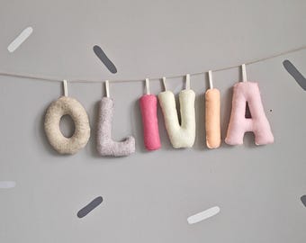 May include: A colourful felt banner spelling "OLIVIA" in pastel shades of beige, lilac, pink, white, and peach. The letters are suspended on a string against a grey wall decorated with scattered, elongated shapes.