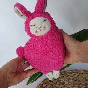 May include: A pink plush toy shaped like a llama with embroidered eyes and a white face. The toy has long, fluffy ears and is being held by two hands.