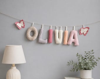 May include: A decorative garland featuring plush, fabric-covered letters spelling the name OLIVIA in shades of beige, lavender, pink, cream, and peach, accented by two felt butterfly ornaments, all suspended from a thin string against a gray wall.