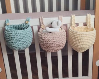 Op de afbeelding: Drie hangende gehaakte manden in lichtblauw, oudroze en lichtgeel, elk voorzien van twee riemen bevestigd met kleine houten knopen. De roze mand bevat een kleine witte pluche beer. De manden hangen aan de spijlen van een wit ledikant.