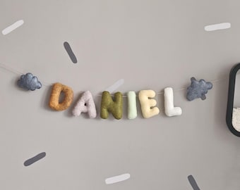May include: A decorative garland featuring soft, plush, felt-like letters spelling the name DANIEL in various colors including mustard, light gray, olive green, pale green, cream, and white, flanked by two small, dark gray cloud-shaped ornaments.