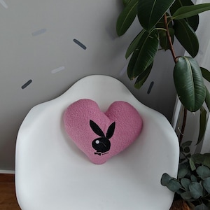 Könnte beinhalten: Ein rosa herzförmiges Kissen mit einem schwarzen Hasenlogo auf einem weißen Stuhl. Der Stuhl hat Holzbeine. Eine große grüne Pflanze befindet sich im Hintergrund, zusammen mit einer grauen Wand mit abstrakten Formen.