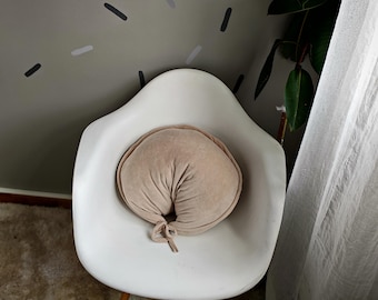 May include: A white, mid-century modern style chair with wooden legs. A round, tan-colored pillow with a knotted tie sits on the chair. The chair is in a room with a neutral color scheme and a plant.