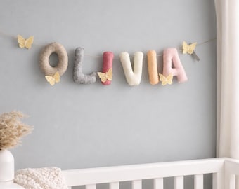 May include: A felt letter garland spelling the name OLIVIA in various soft colors including grey, pink, and cream. The letters are strung together on a thin cord and decorated with small, delicate, yellow lace butterfly cutouts.