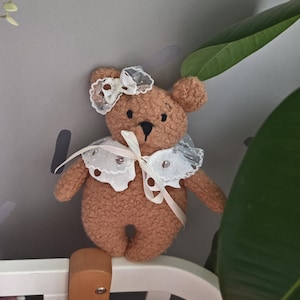 Könnte beinhalten: Ein brauner Teddybär mit weißem Spitzenkragen und Schleife. Der Bär hat schwarze Knopfaugen und eine kleine schwarze Nase. Die Schleife ist ebenfalls aus weißer Spitze. Der Bär steht auf einem weißen Kinderbett.
