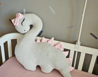 May include: A soft, light gray dinosaur-shaped plush pillow featuring a long, curved neck, a pink bow attached to the head, and a row of pink triangular spikes along the back. The dinosaur has embroidered closed eyes and a small mouth.