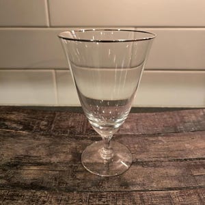 May include: A clear glass goblet with a silver rim. The goblet has a tall, slender stem and a round base.