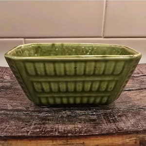 May include: A square, green ceramic bowl with a textured, basket-weave design. The bowl has a slightly irregular shape and sits on a wooden surface. The bowl is a muted green color.