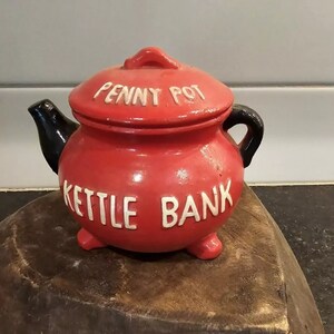May include: Red ceramic kettle-shaped piggy bank with a black lid and handle. The bank has white lettering that reads "Penny Pot Kettle Bank".