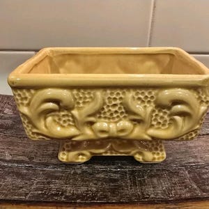 May include: A rectangular, yellow ceramic planter with ornate, raised floral and scrollwork designs. The planter has a square base and is elevated on four small feet. The interior is a lighter shade of yellow.