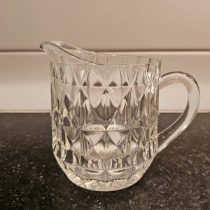 May include: A clear glass creamer with a diamond pattern. The creamer has a handle and a spout.