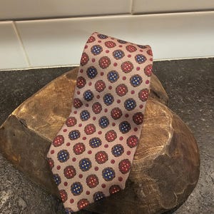 May include: A patterned tie with a light brown background and repeating red and blue floral designs. The tie is made of a smooth fabric and is displayed on a wooden surface. The tie is a classic accessory for formal or business attire.