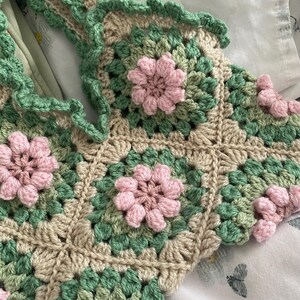 以下が含まれることがあります： 緑とベージュのグラニー・スクエア模様のクロシェトップ。ピンクの花のアクセントが施されています。