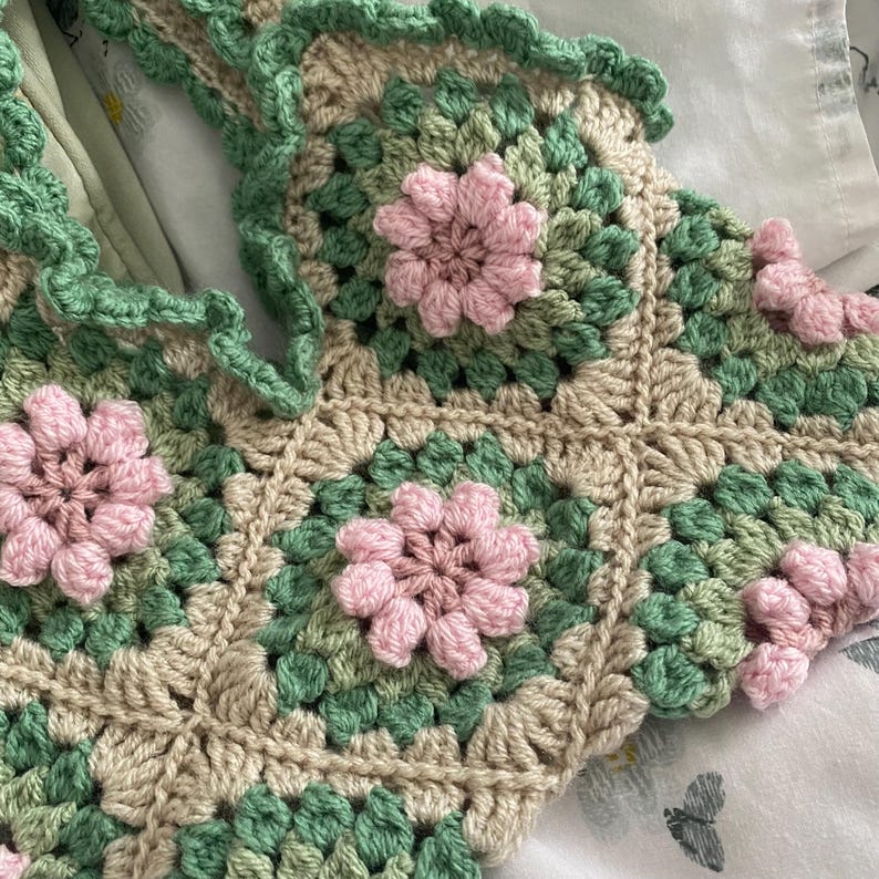 以下が含まれることがあります： 緑とベージュのグラニー・スクエア模様のクロシェトップ。ピンクの花のアクセントが施されています。