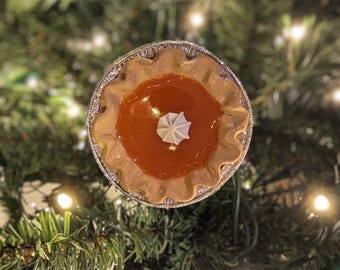 Peut inclure: Une petite tarte avec une croûte dorée et une garniture orange brillante, surmontée d'une touche de crème fouettée blanche. La tarte est dans un récipient en papier d'aluminium, sur un fond de branches de sapin vertes et de lumières scintillantes.