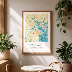 May include: A framed wall art print featuring a watercolor map with a red heart icon marking a specific location. Below the map, the text reads John and Emily, WHERE IT ALL BEGAN, and the date 7/18/2024 in a clean, black sans-serif font.