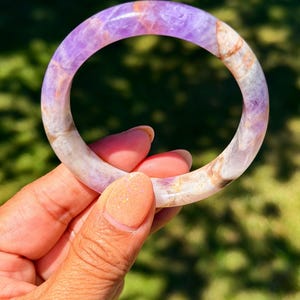 May include: A smooth, circular bangle bracelet crafted from polished stone, featuring a marbled pattern of vibrant purple amethyst hues blended with translucent white and warm tan inclusions throughout the entire rounded surface of the jewelry.