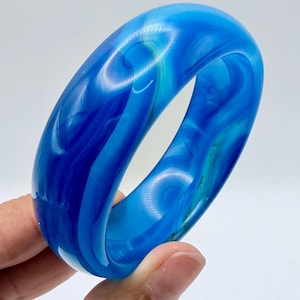 May include: A thick, polished bangle bracelet featuring a vibrant, translucent blue color with swirling patterns of lighter and darker shades of blue throughout the smooth, rounded surface of the circular accessory.