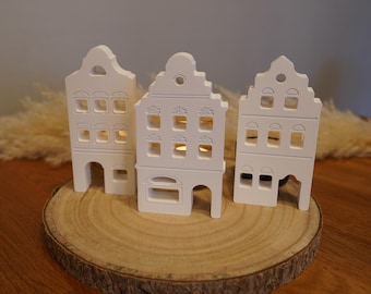 Peut inclure: Trois bougeoirs blancs en forme de maison avec des fenêtres et des portes découpées, disposés sur une tranche de bois ronde. Les bougeoirs sont éclairés de l'intérieur, créant une lueur chaleureuse. Les maisons ont un design architectural traditionnel.