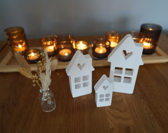 Peut inclure: Trois maisons en bois blanc avec des fenêtres et des cœurs découpés, disposées en rangée sur une table en bois. Les maisons sont éclairées par des bougies dans des bougeoirs en verre derrière elles.