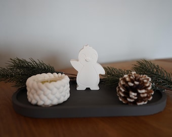 Peut inclure: Une figurine de pingouin en céramique blanche avec un visage souriant, un bougeoir en céramique blanche en forme de panier et une pomme de pin brune sur un plateau gris.