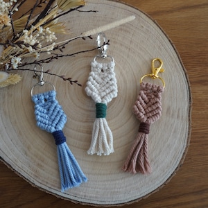 May include: Three macrame keychains with tassels. The keychains are in blue, white, and brown with green and brown accents. The keychains are hanging from a wooden surface.