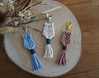 Peut inclure: Trois porte-clés en macramé avec des glands. Les porte-clés sont en bleu, blanc et marron avec des accents verts et marrons. Les porte-clés sont suspendus à une surface en bois.