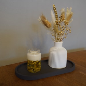 May include: A white ceramic vase with a ribbed texture holds a bouquet of dried flowers with white and gold accents. A clear glass jar with a white candle and green and gold accents sits on a gray oval tray.