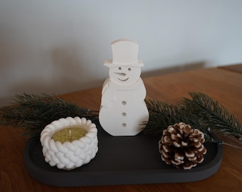 Peut inclure: Bougie en forme de bonhomme de neige en céramique blanche avec un plateau noir et deux branches de pin avec une pomme de pin.