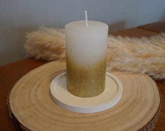 Peut inclure: Une bougie blanche et dorée à paillettes avec une mèche, posée sur un petit plateau circulaire blanc. La bougie a un effet dégradé, passant du blanc aux paillettes dorées. La bougie et le plateau sont sur une tranche de bois.