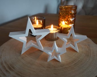 Peut inclure: Quatre étoiles en bois blanc de tailles différentes sont disposées sur une tranche de bois. Trois bougeoirs en verre avec des bougies allumées sont placés derrière les étoiles.