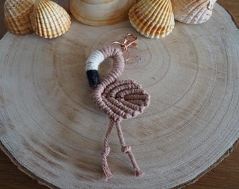 Peut inclure: Un porte-clés en macramé en forme de flamant rose et blanc avec des accents noirs. Le porte-clés est suspendu à une surface en bois.