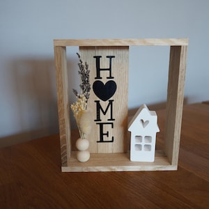 May include: Wooden framed decorative piece with the word "HOME" in black letters and a black heart. It includes a small white house with a heart cutout and a vase with dried flowers. The frame is made of light-colored wood.