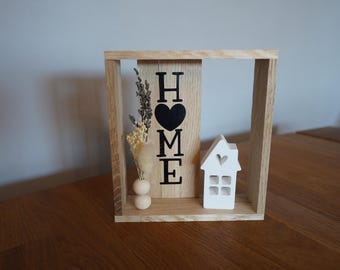 Peut inclure: Cadre décoratif en bois clair avec le mot « HOME » en lettres noires et un cœur noir. Il comprend une petite maison blanche avec une découpe en forme de cœur et un vase avec des fleurs séchées.