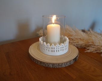 Peut inclure: Une bougie blanche allumée dans une lanterne en verre transparent, posée sur une base décorative blanche avec une silhouette de ville. La lampe repose sur une tranche de bois ronde et naturelle. La scène est sur une table en bois.