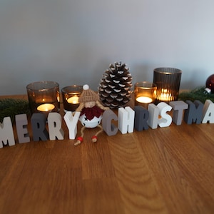 May include: A festive Christmas display on a wooden surface. The arrangement includes lit candles in glass holders, decorative ornaments, a pine cone, and the words "MERRY CHRISTMAS" spelled out in wooden letters. A small doll is also included.