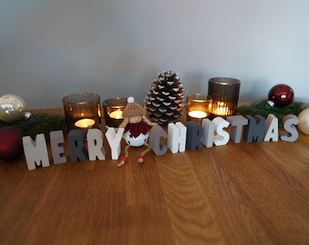 Peut inclure: Une composition de Noël sur une surface en bois. Elle comprend des bougies allumées dans des photophores, des boules décoratives, une pomme de pin et l'inscription "MERRY CHRISTMAS" en lettres de bois. Une petite poupée est également présente.