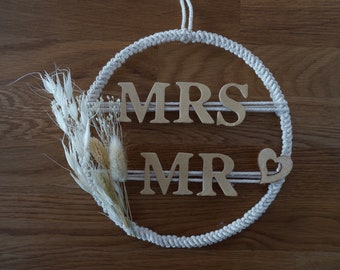 Peut inclure: Une couronne en corde blanche avec les mots "MRS" et "MR" en lettres de bois. Un petit cœur en bois est attaché au bas de la couronne. Des fleurs blanches séchées sont attachées à gauche de la couronne.