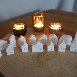 May include: A collection of small white houses with detailed designs, arranged on a wooden slice. The houses are illuminated by candlelight from three glass candle holders.