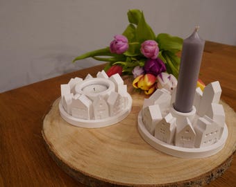 Peut inclure: Deux bougeoirs circulaires blancs présentant un design de village miniature avec de petites structures de maisons sur le pourtour. L'un contient une bougie chauffe-plat, tandis que l'autre porte une bougie grise haute et effilée.