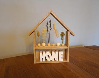 Peut inclure: Étagère en bois en forme de maison avec le mot "HOME" en lettres blanches. L'étagère contient des fleurs séchées et des sphères décoratives en bois. La structure est en bois clair et repose sur une surface en bois.