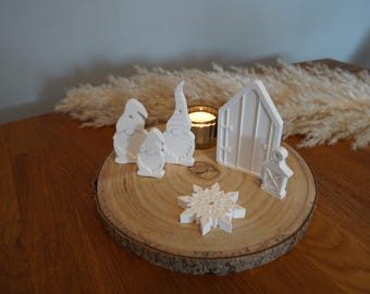 Peut inclure: Ensemble de figurines décoratives blanches, comprenant des gnomes, une maison, une lanterne et un flocon de neige, disposées sur une tranche de bois. Une bougie allumée dans un support doré ajoute de la chaleur. Sur une table en bois.