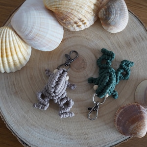 Könnte beinhalten: Zwei Makramee-Frosch-Schlüsselanhänger, einer grau und einer türkis, mit Holzperlen-Akzenten und silberfarbenen Verschlüssen. Die Schlüsselanhänger werden auf einer Holzscheibe mit Muscheln präsentiert.