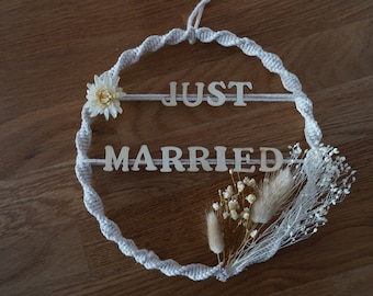 Peut inclure: Une couronne en macramé blanche avec les mots "JUST MARRIED" en lettres de bois. La couronne est décorée de fleurs séchées et de verdure.