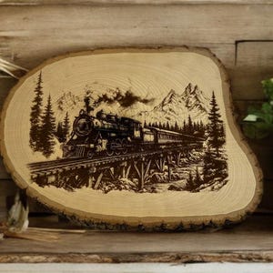 May include: A wood slice with a detailed engraving of a vintage train crossing a trestle bridge, set against a backdrop of mountains and pine trees. The artwork is in shades of brown and black.