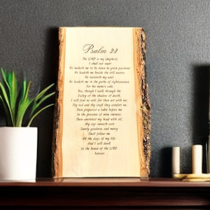 May include: A wooden plaque with the text of Psalm 23, displayed on a dark wooden shelf. A small potted plant, a white sphere, and a gold-framed mirror are also on the shelf. The plaque has a natural bark edge.