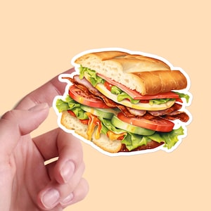 Könnte beinhalten: Ein großes Sandwich mit verschiedenen Füllungen, darunter Salat, Tomate, Gurke, Käse und Fleisch. Das Sandwich ist auf einem weißen Brötchen und wird von einer Hand gehalten.
