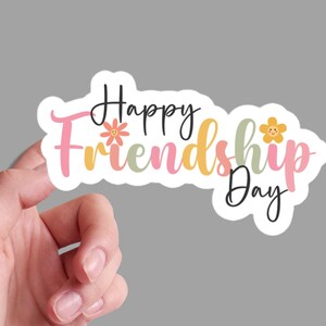 May include: A white sticker with the text "Happy Friendship Day!" in pink, yellow, and green lettering. The text is surrounded by two yellow flowers with smiley faces.