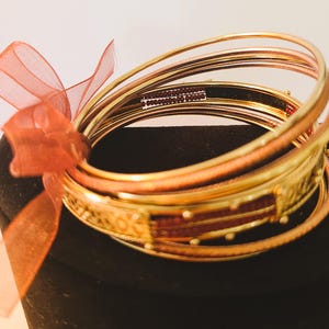 May include: A set of gold-toned metal bangles featuring a mix of smooth and textured surfaces. One bangle includes a rectangular panel with small dark red beads and a sheer orange ribbon tied to the side of the stack of circular bracelets.